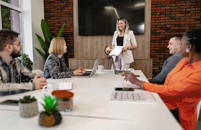 A photo of a group of people sitting around a table with laptops and note, one person is standing at the head of the table