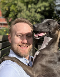 Adam Richardson smiling before a leafy tree with a dog with its mouth open and tongue out. 