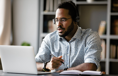 A person sitting at a desk wearing headphones, with a pen, paper and a laptop