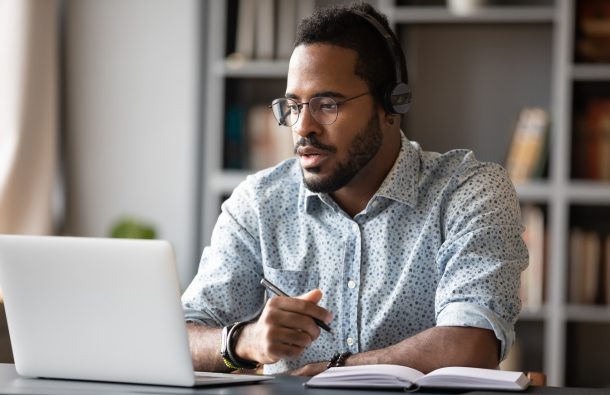 A person sitting at a desk wearing headphones, with a pen, paper and a laptop
