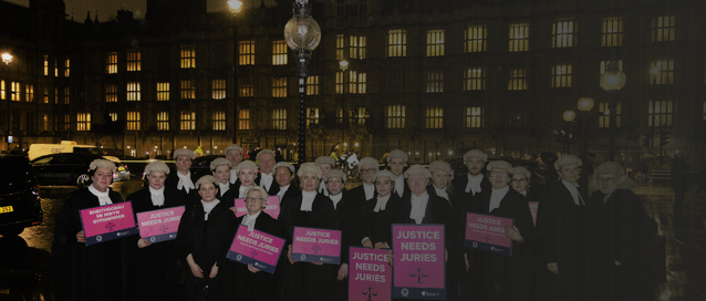 A photo of a group of barristers in wigs and gowns holding signs saying 'Justice needs juries', in front of the Houses of Parliament