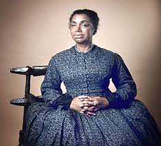 Photograph of a Black woman sitting in a wooden chair looking at the camera with her hands folded on her knees. She is wearing a blue patterned dress in a late 18th/early 19th century style. 