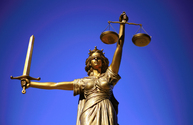 A photo of a statue of lady justice, holding a sword out to one side and a set of scales in front with a blindfold on, in front of a blue background