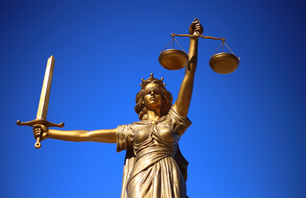 A photo of a statue of lady justice, holding a sword out to one side and a set of scales in front with a blindfold on, in front of a blue background