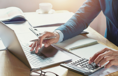A photo of someone at a desk with a laptop, calculator, open pad and a pen
