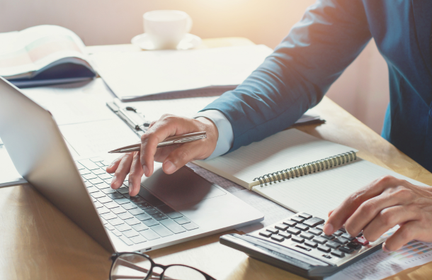 A photo of someone at a desk with a laptop, calculator, open pad and a pen