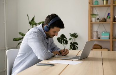 A person with headphones on, sitting at a table with a laptop open, writing notes in a notepad  
