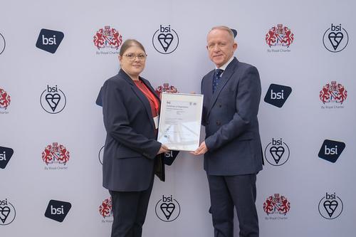 A woman in a blue suit holds a certificate award with a man in a blue suit and spotty tie. Facilities manager Chiara Begg and head of facilities Andy Curtis with the ISO 14001 accreditation award
