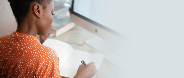 A woman sitting at a desk writing in a notepad in front of a computer screen with long text on it.