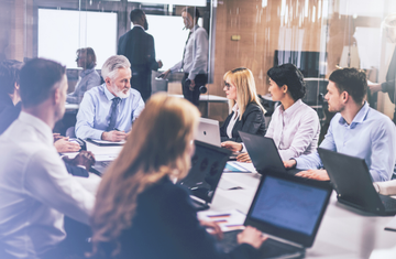 A group of people sitting around a table with laptops in front of them, all looking at one person who is talking 